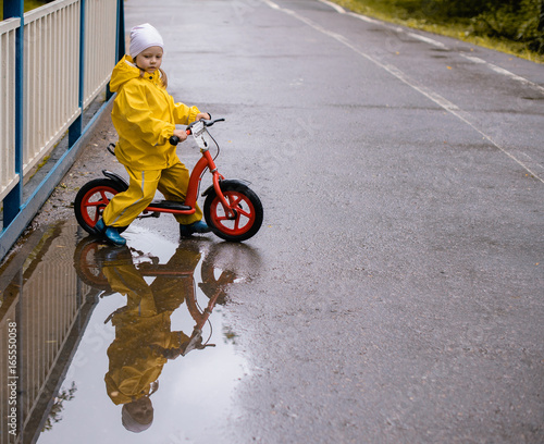 Billede på lærred little girl in a autumn park on a bike near a puddle