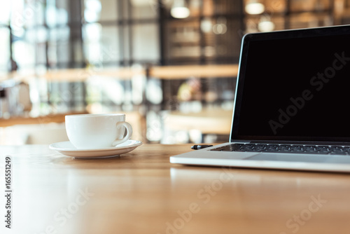 close up view of cup of coffee and laptop with blank screen on wooden table in cafe