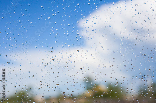 Rain drops on glass window next to blue sky and blurred green trees and white clouds. Summer weather concept.