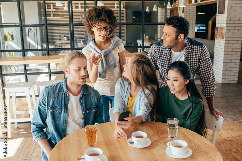 Young happy multiethnic friends looking at smartphone while resting in cafe Young happy multiethnic friends looking at smartphone while resting in cafe