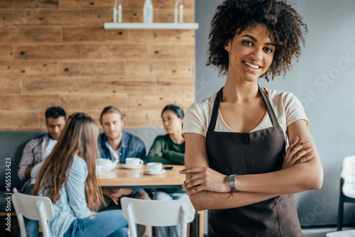 Smiling african american waitress standing with customers sitting behind in cafe