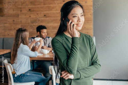Asian woman talking on smartphone while her friends sitting behind in cafe