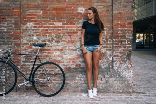 An outdoor portrait of a young cute student girl wearing black blank t-shirt and blue jeans shorts with a fixed gear bicycle while standing on the brick wall background. Empty space for text or design