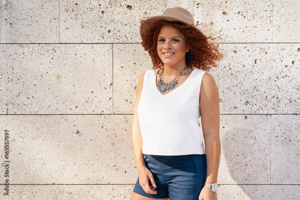 A red-haired young girl wearing casual clothes and hat is looking at camera while standing beside the light gray wall. Young woman with freckle face is standing on the wall background. A red-haired young girl wearing casual clothes and hat is looking at camera while standing beside the light gray wall. Young woman with freckle face is standing on the wall background.