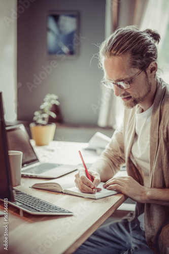 Side view of freelance man sitting at wooden table writing in notepad. Man at home office working making notes.