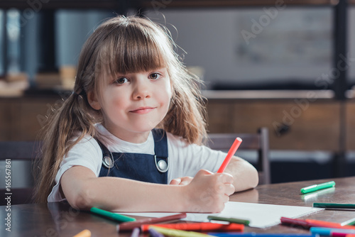 portrait of cute smiling girl sitting at table and drawing picture at home