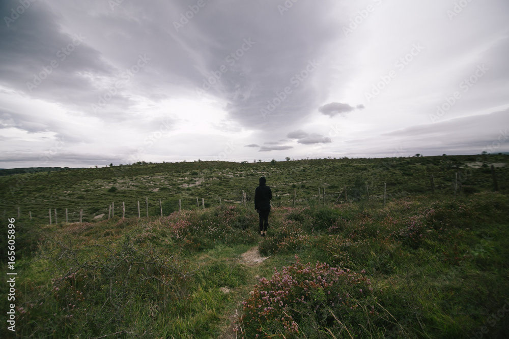 Fototapeta premium Silhouette of lonely traveler in black outfit standing in middle of field or meadow on grey rainy day with sad atmosphere of loneliness and doom
