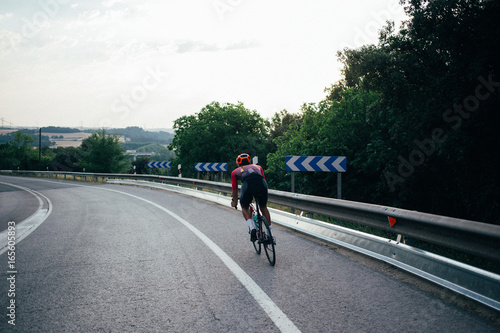 Fototapeta Naklejka Na Ścianę i Meble -  Fit healthy sportsman rides carbon aero bicycle on empty sunset road with no traffic, beautiful scenery around him, unconventional vacation ideas