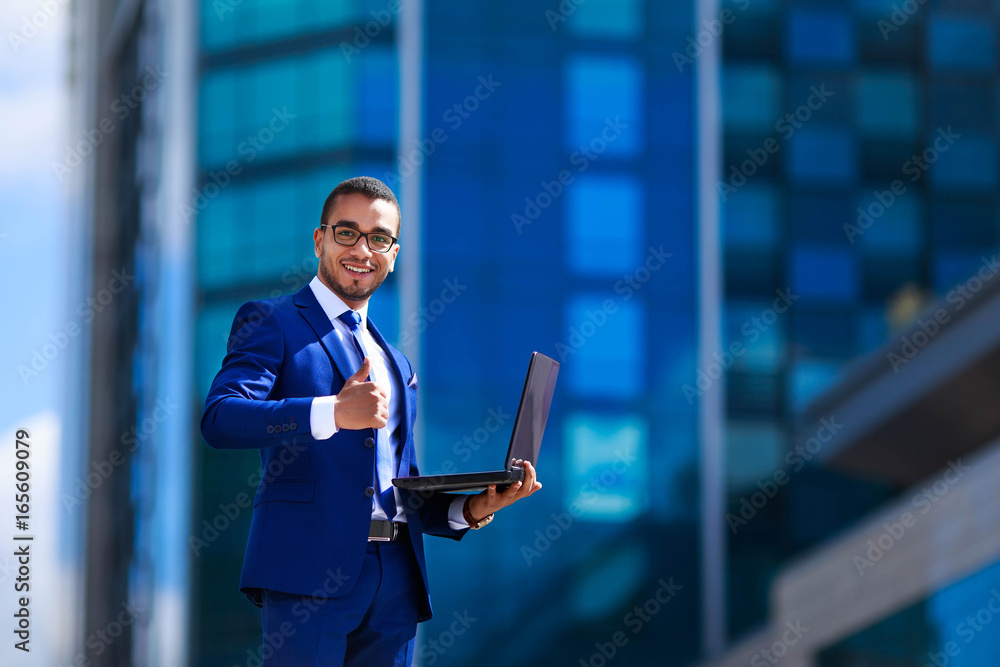 Business man in suit holding laptop while standing near office building,demonstrating thumb up gesture, expressing success Business man in suit holding laptop while standing near office building,demonstrating thumb up gesture, expressing success