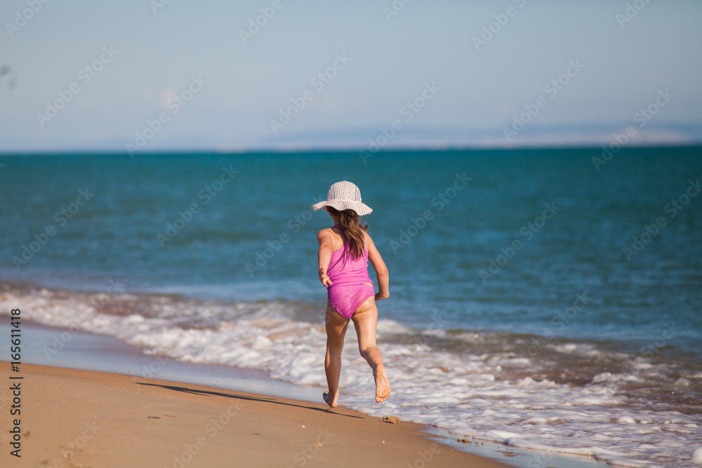 young girl in swimsuit and hat runs along the line of surf on a sandy beach