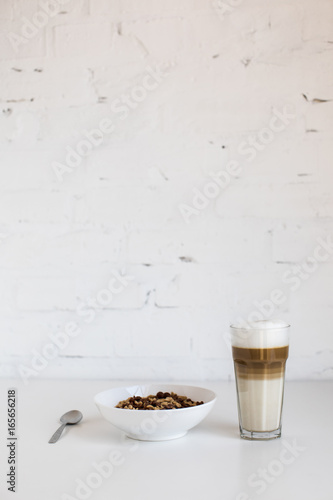 glass of coffee latte and cornflakes in bowl with spoon on white table