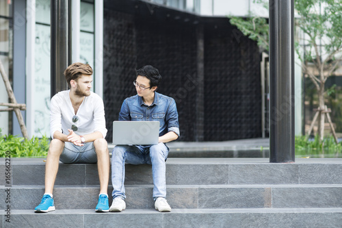 Two asian man and caucasian man are sitting, talking, meeting, discussing and working with computer laptop at stair outdoor college.