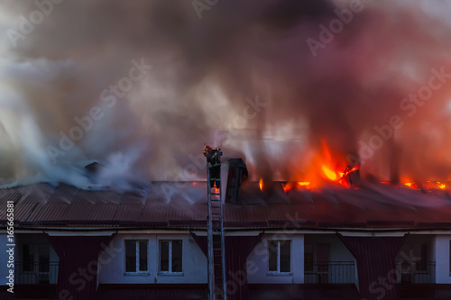 Burning fire flame with smoke on the apartment house roof in the city, firefighter or fireman on the ladder extinguishes fire