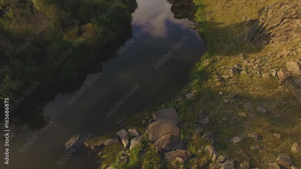 AERIAL: A camera rises from the river to the village among a beautiful summer landscape with rocks, trees and green meadow during sunset. AERIAL: A camera rises from the river to the village among a beautiful summer landscape with rocks, trees and green meadow during sunset.