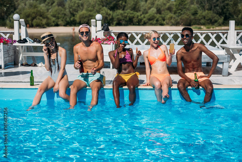 Group of young multiethnic friends sitting with beverages at poolside during vacation at resort