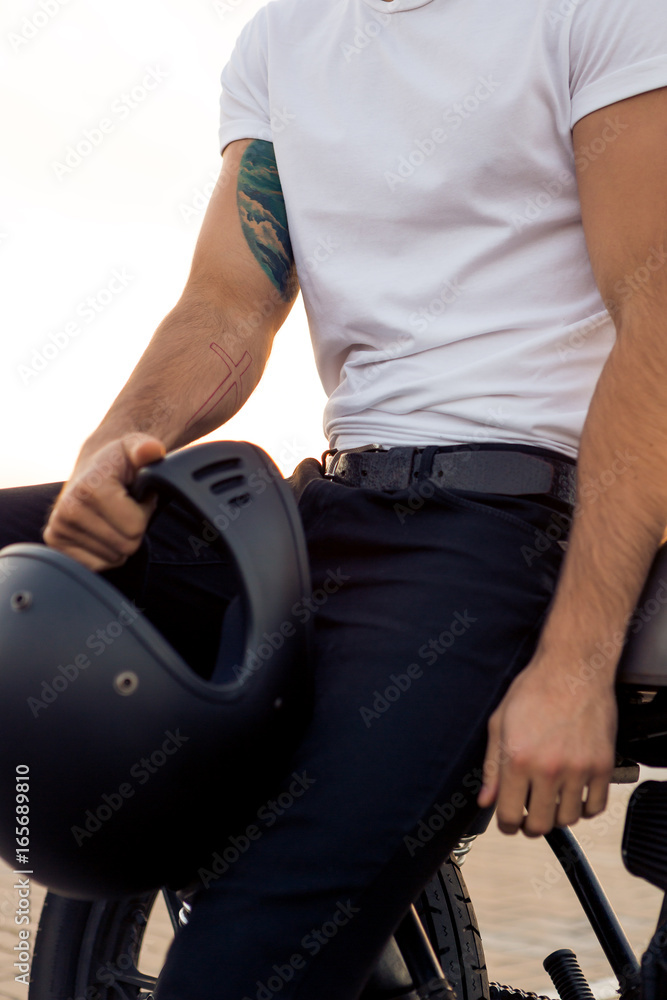 Fototapeta premium Close up of a handsome rider male in white blank t-shirt with helmet near classic style cafe racer motorcycle at sunset. Bike custom made in vintage garage. Brutal urban lifestyle. Outdoor portrait.