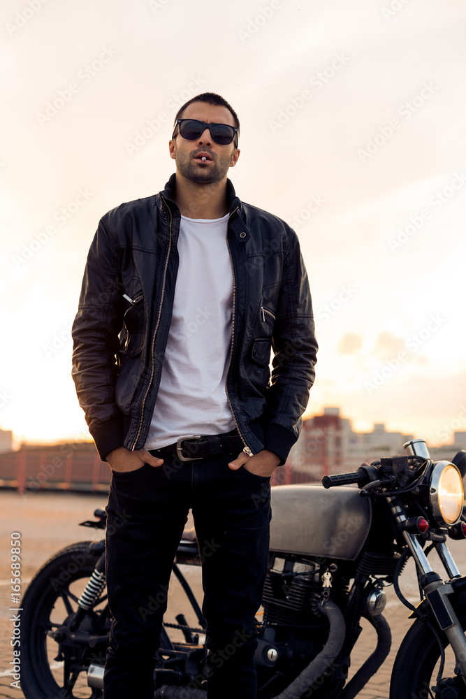Handsome rider man with beard and mustache in black biker jacket, white t-shirt and fashion sunglasses smoking cigaret near classic style cafe racer motorbike at sunset. Brutal fun urban lifestyle.