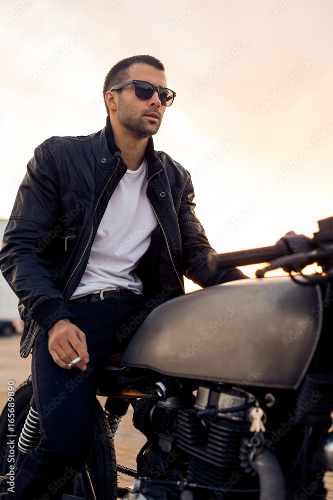 Beautiful happy rider man with beard and mustache in black biker jacket,  jeans and fashion sunglasses smoking cigaret sit on classic style cafe racer  motorbike at sunset. Brutal fun urban lifestyle. Photos