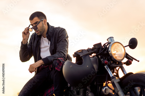 Handsome rider man with beard and mustache in black biker jacket take off sunglasses on classic style cafe racer motorcycle at sunset. Bike custom made in vintage garage. Brutal fun urban lifestyle.
