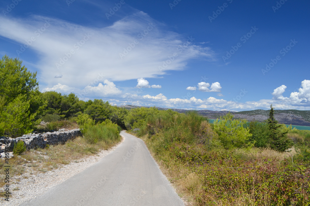 Asphalt road in Western part of Vransko jezero Nature Reserve, green trees and blue sky with clouds, Dalmatia, Croatia Asphalt road in Western part of Vransko jezero Nature Reserve, green trees and blue sky with clouds, Dalmatia, Croatia