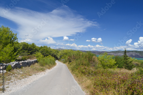 Asphalt road in Western part of Vransko jezero Nature Reserve, green trees and blue sky with clouds, Dalmatia, Croatia