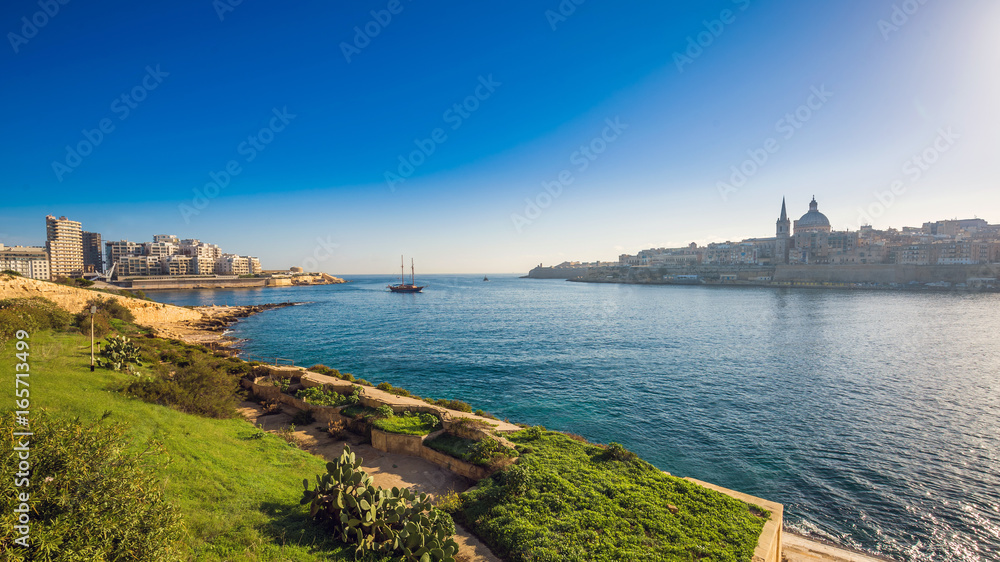 Valletta, Malta - Skyline view of the ancient city of Valletta and Sliema at sunrise shot from Manoel island at spring time with sailing boat, St.Paul's cathedral, blue sky and green grass