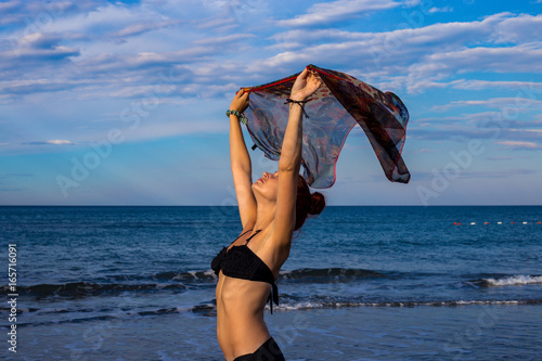 Slim good looking tanned redhead with arms outstretched holding scarf while wind is blowing on the beach in black bikini with blue sea and sky in the background during summer