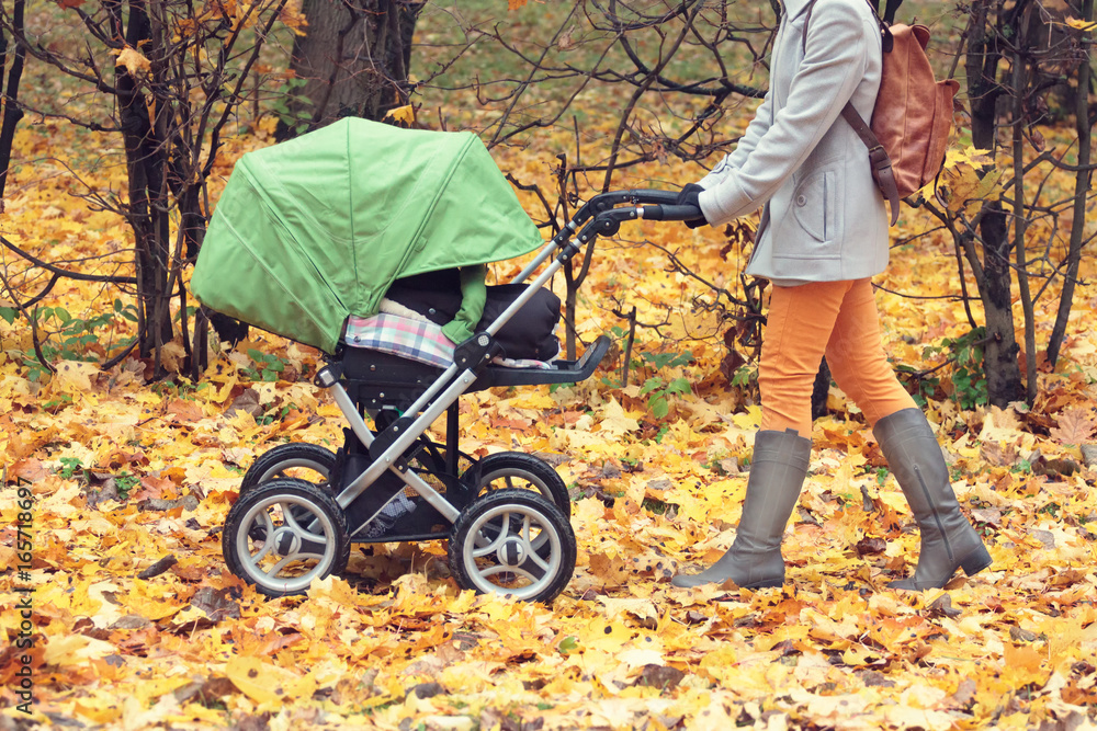 Fototapeta premium Mother pushing stroller in autumn park. Young woman strolling with a baby in buggy outdoors. Dry orange leaves on the ground