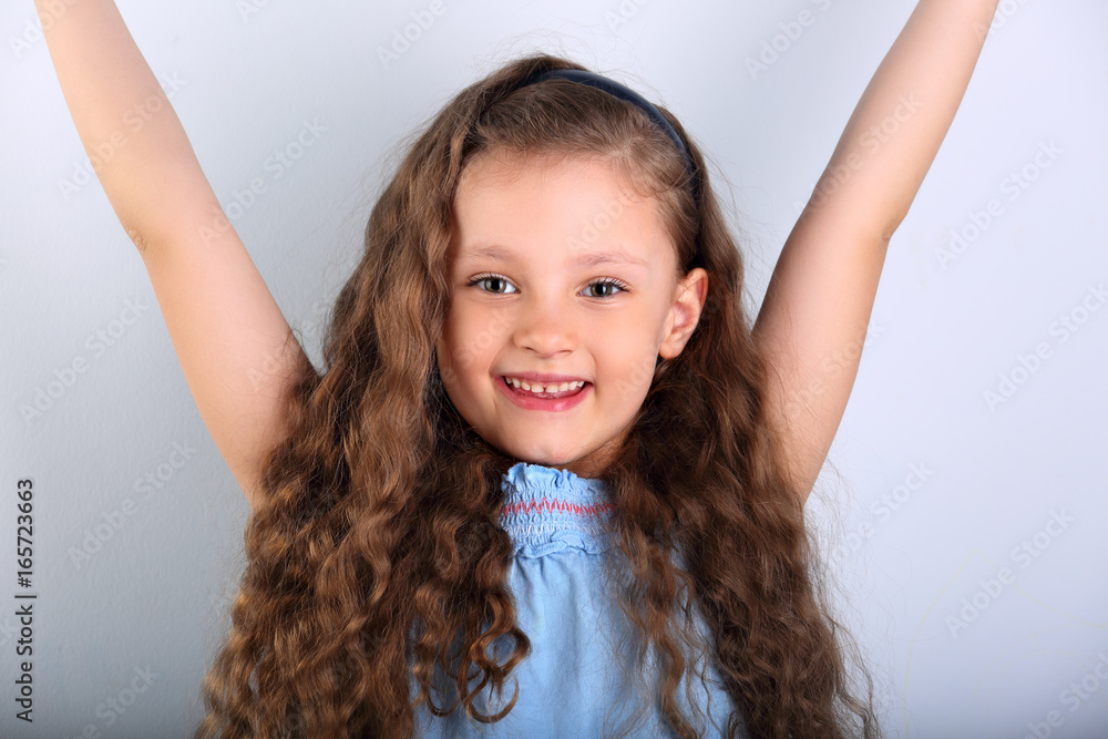 Beautiful smiling natural joying kid girl looking with long curly hair style and rises her hands up on blue background. Closeup portrait