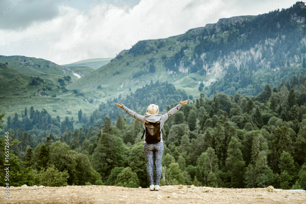 The girl stands with her back with a backpack on top of the mountain against the background of the Alpine mountains and the heavenly landscape. Travel and tourism in the open air.
