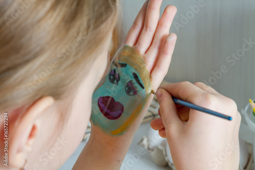 Девочка рисует лицо акварельными красками на своей ладони.