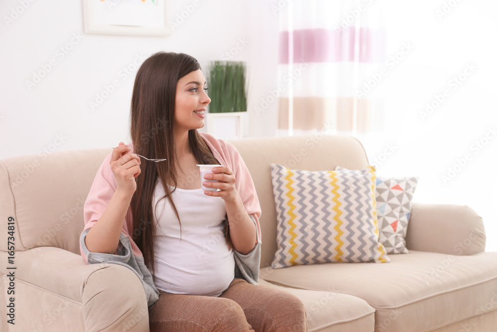 Young pregnant woman sitting on sofa and eating yogurt in light room Young pregnant woman sitting on sofa and eating yogurt in light room