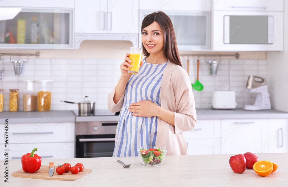 Young pregnant woman drinking juice while standing near table in kitchen