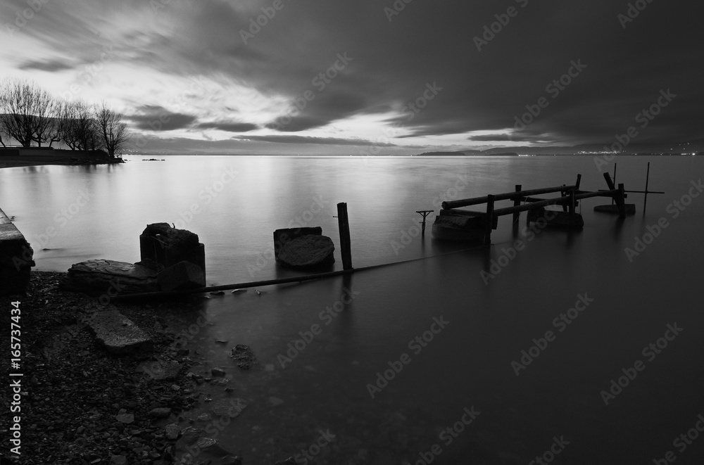 Fototapeta premium A pier built of wood and stones on a lake at dusk, with perfectly still water and moving clouds