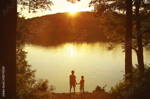 Children at the water watching the setting sun. Two boys holding hands standing on the shore. Back view. Copy space for your text  photo