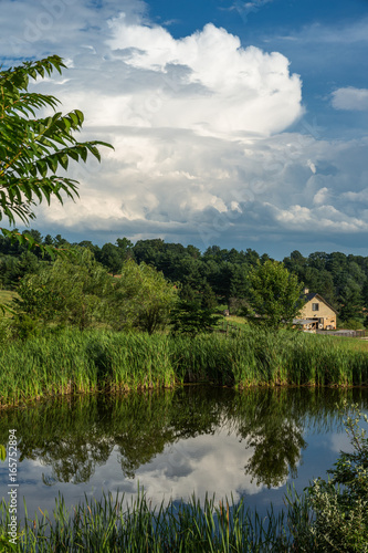 A small, reed-lined pond reflects the gathering storm in the background on a sleepy horse farm in the countryside.
