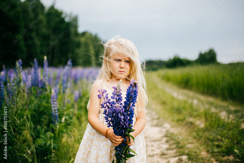 Lost, lonely and beautiful baby kid little blonde girl with offended pathetic poor tear-stained face standing on field with flowers road at nature outdoor in white dress and holding bouquet of lupines Lost, lonely and beautiful baby kid little blonde girl with offended pathetic poor tear-stained face standing on field with flowers road at nature outdoor in white dress and holding bouquet of lupines