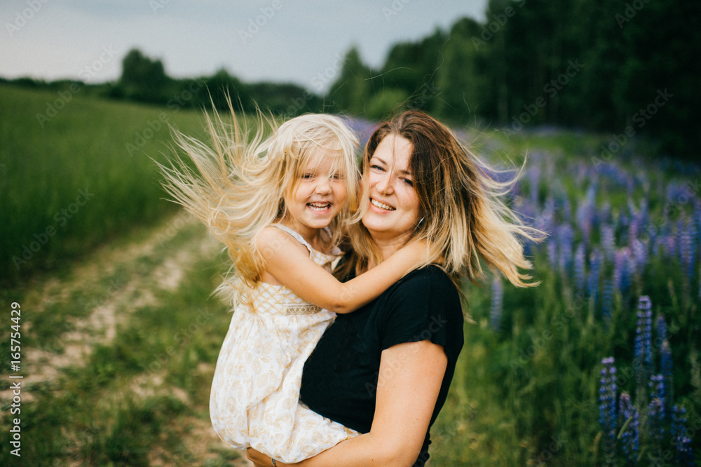Fototapeta premium Young mother with long hair holding little daughter princess at hands. Playing with child. Pure, happy, kind, expressive emotional faces. Family at nature. Artistic kid. Laughing, smiling, embracing.