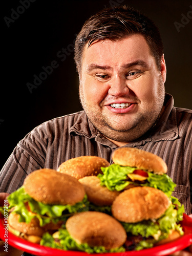 Fat man eating fast food hamberger and carries treat for friends on tray. Breakfast for overweight person. Poor food leads to obesity. Person regularly overeats concept on black background.