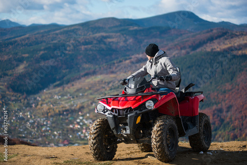 Man in winter clothes on a red quad bike on a mountain top enjoying the view of the mighty mountains behind him. Blurred background of mountain scenery