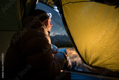 View from inside a tent on the male tourist have a rest in his camping at night. Man with a headlamp sitting in the tent near campfire under beautiful night sky full of stars and milky way