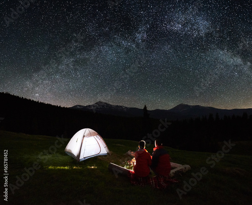 Night camping. Silhouette of couple tourists sitting at a campfire near illuminated tent under beautiful night sky full of stars and milky way. Astrophotography