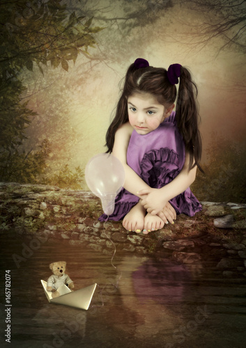 A little girl with a pensive look, sitting on a rock above the water and a paper boat 