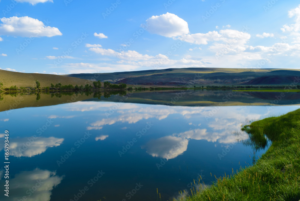 Landscape of calm lake, hills, green grass and blue sky in Altai mountains. White clouds reflected in water. Chuya prairie, Altay Republic, Siberia, Russia.