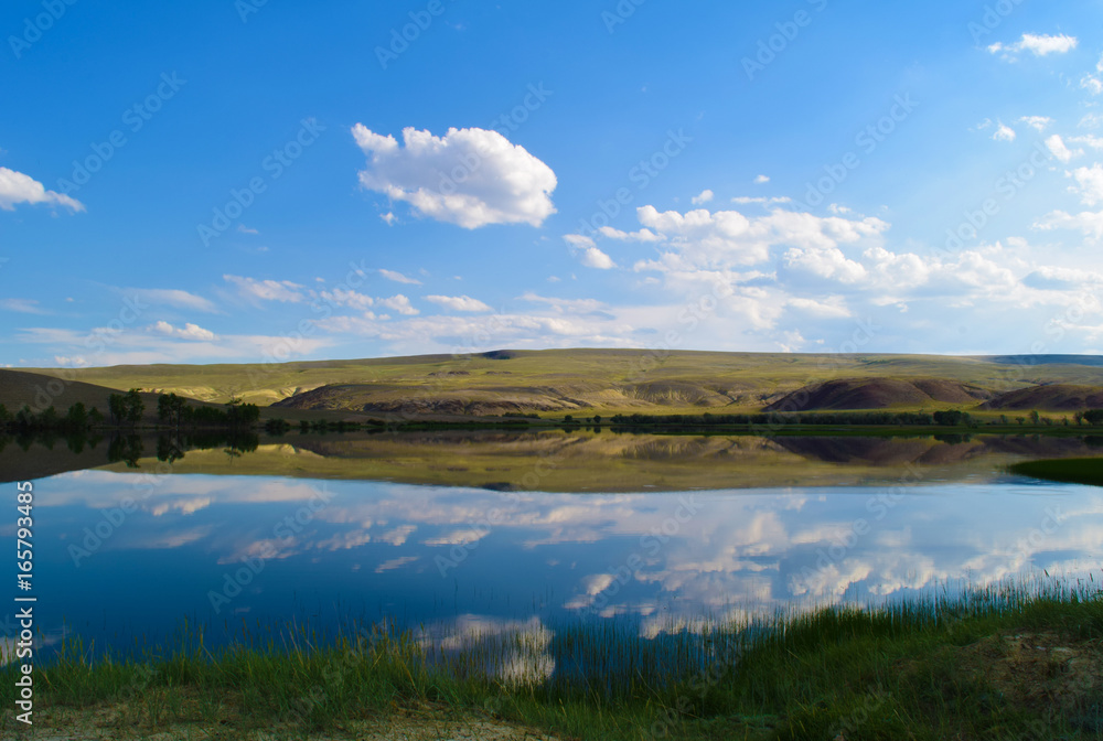 Landscape of calm lake, hills, green grass and blue sky in Altai mountains. White clouds reflected in water. Chuya steppe, Altay Republic, Siberia, Russia.