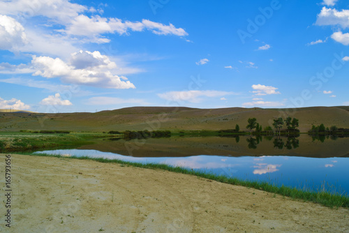 Landscape of calm lake, hills, dirt road and blue sky in Altai mountains. White clouds reflected in water. Chuya steppe, Altay Republic, Siberia, Russia.