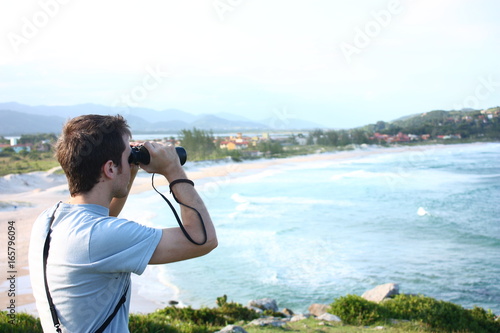 Young man near the beach looking away through binoculars on the top of a hill. 