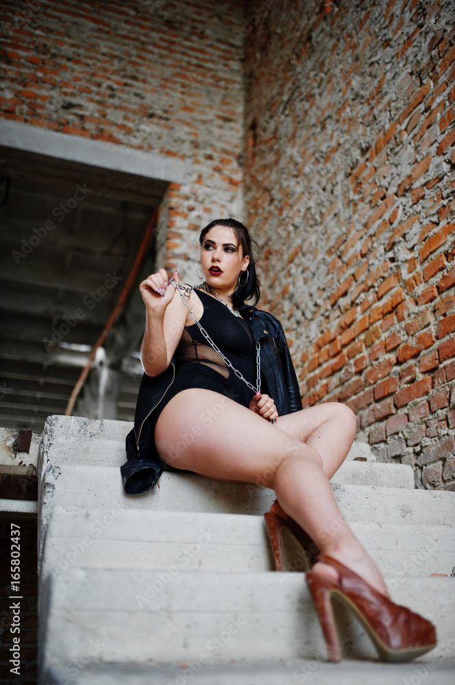 Brunette plus size sexy woman, wear at black one piece and leather jacket,with chain at abadoned place sitting on stairs. BDSM theme.