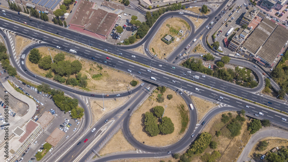 Vista aerea di un uscita autostradale a roma. Le auto corrono veloci lungo l'autostrada mentra alcune si stanno immettendo o ne sta uscendo tramite la corsia di accellerazione o decellerazione.