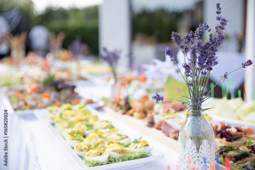 On a white table appetizing appetizers of stuffed eggs, vegetables, different kinds of sandwiches and a beautiful glass vase with blue wildflowers On a white table appetizing appetizers of stuffed eggs, vegetables, different kinds of sandwiches and a beautiful glass vase with blue wildflowers